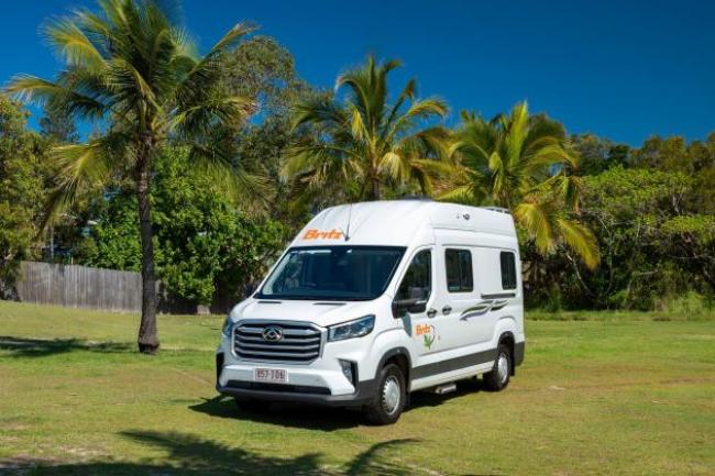 White Britz campervan parked on a grassy field with palm trees under a clear blue sky.