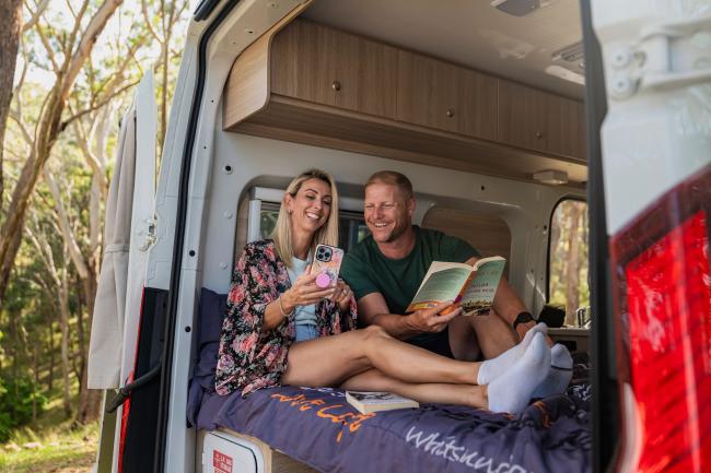 Smiling couple inside a campervan bed; the woman shows her phone to the man holding an open book.