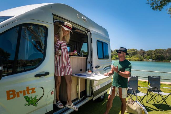 A couple relaxes beside a Britz campervan parked by a river with camping chairs set up on the grass.