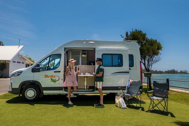 A couple stands beside a white Britz campervan parked on grass near a coastal waterway.