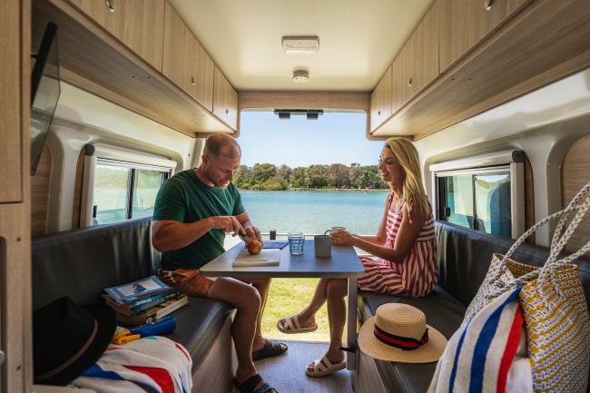 Couple dining at a table inside a campervan, looking out the open back doors at a river.