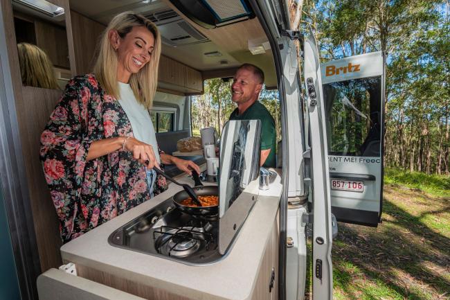 A woman cooks on a stove inside a campervan while a man smiles at her from the open side door.