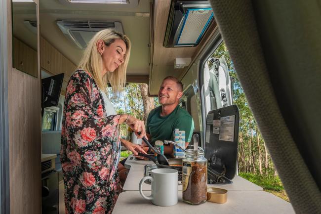 A woman cooks in a campervan kitchen while a man looks on from the open doorway.