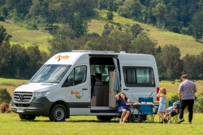 A family relaxes next to a white Britz campervan in a grassy field with rolling hills.