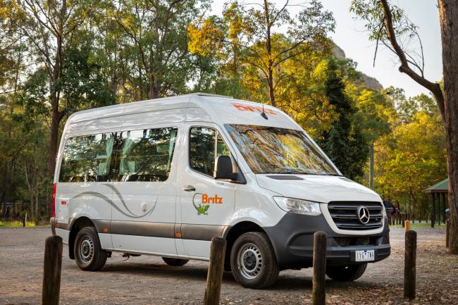 White Britz campervan parked in a tree-lined clearing behind timber posts.