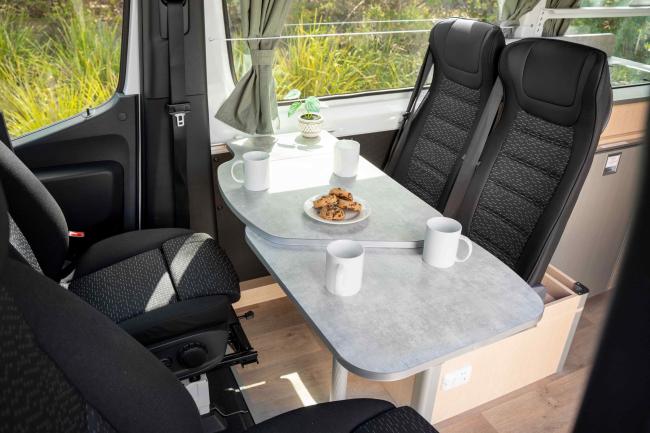 Campervan dining area with four black seats facing a grey table set with mugs and a plate of biscuits.