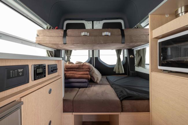 Campervan interior featuring timber cabinetry, built-in microwave, control panel, and a two-tier bed system.