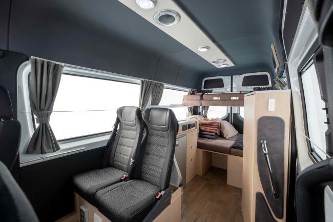 Campervan interior featuring two black passenger seats, light timber cabinetry, and a rear bed setup.