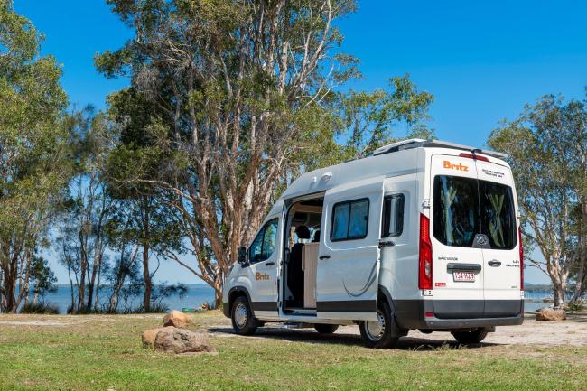 White Britz campervan with an open side door, parked on grass near trees and water under a clear blue sky.