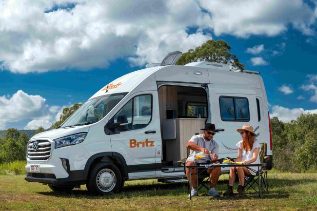 A man and woman sit outside a white Britz campervan in a grassy field, pouring drinks at a camping table.