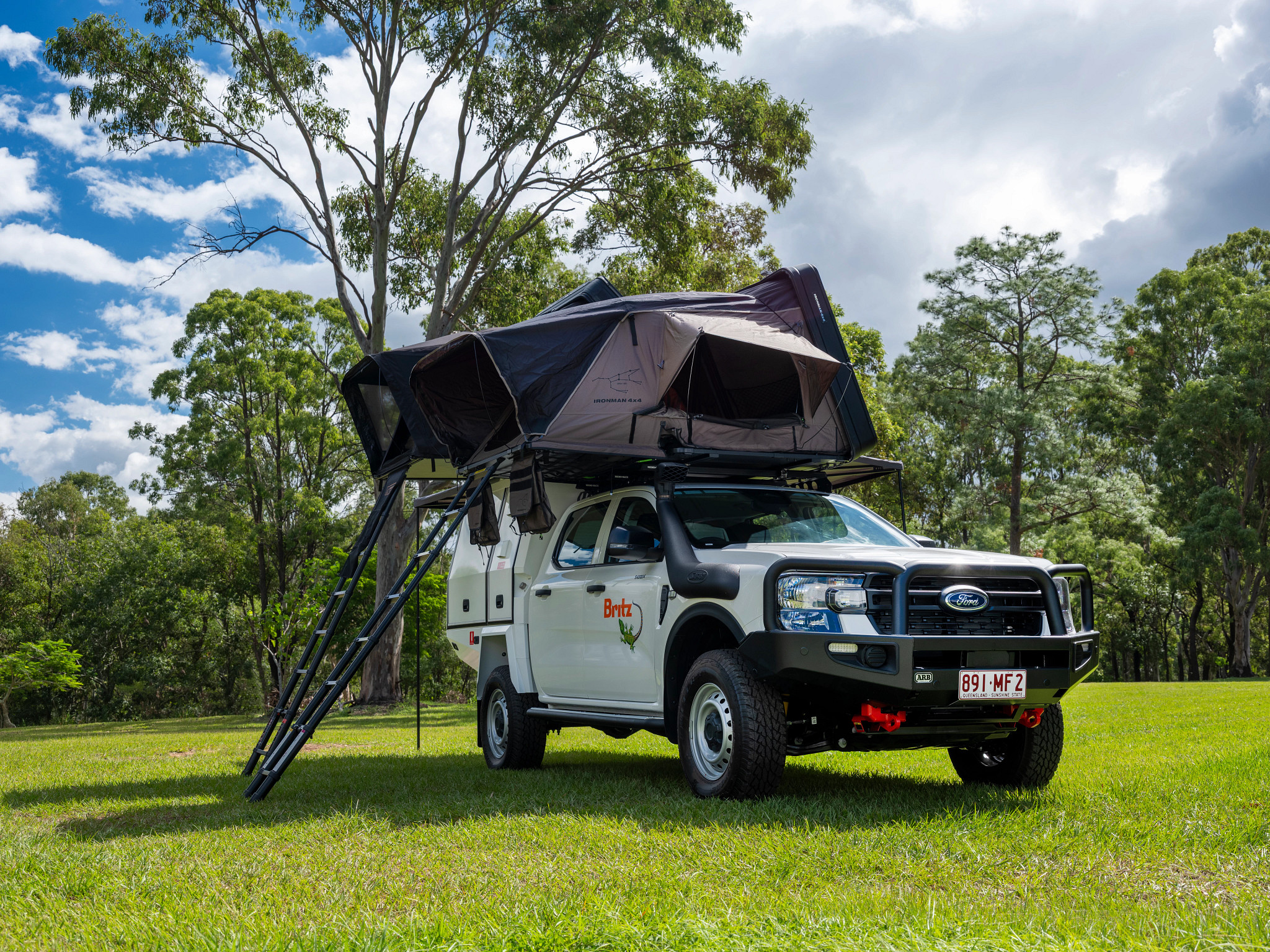 White Britz 4WD Ford campervan with an open rooftop tent and ladder, parked on green grass surrounded by trees.