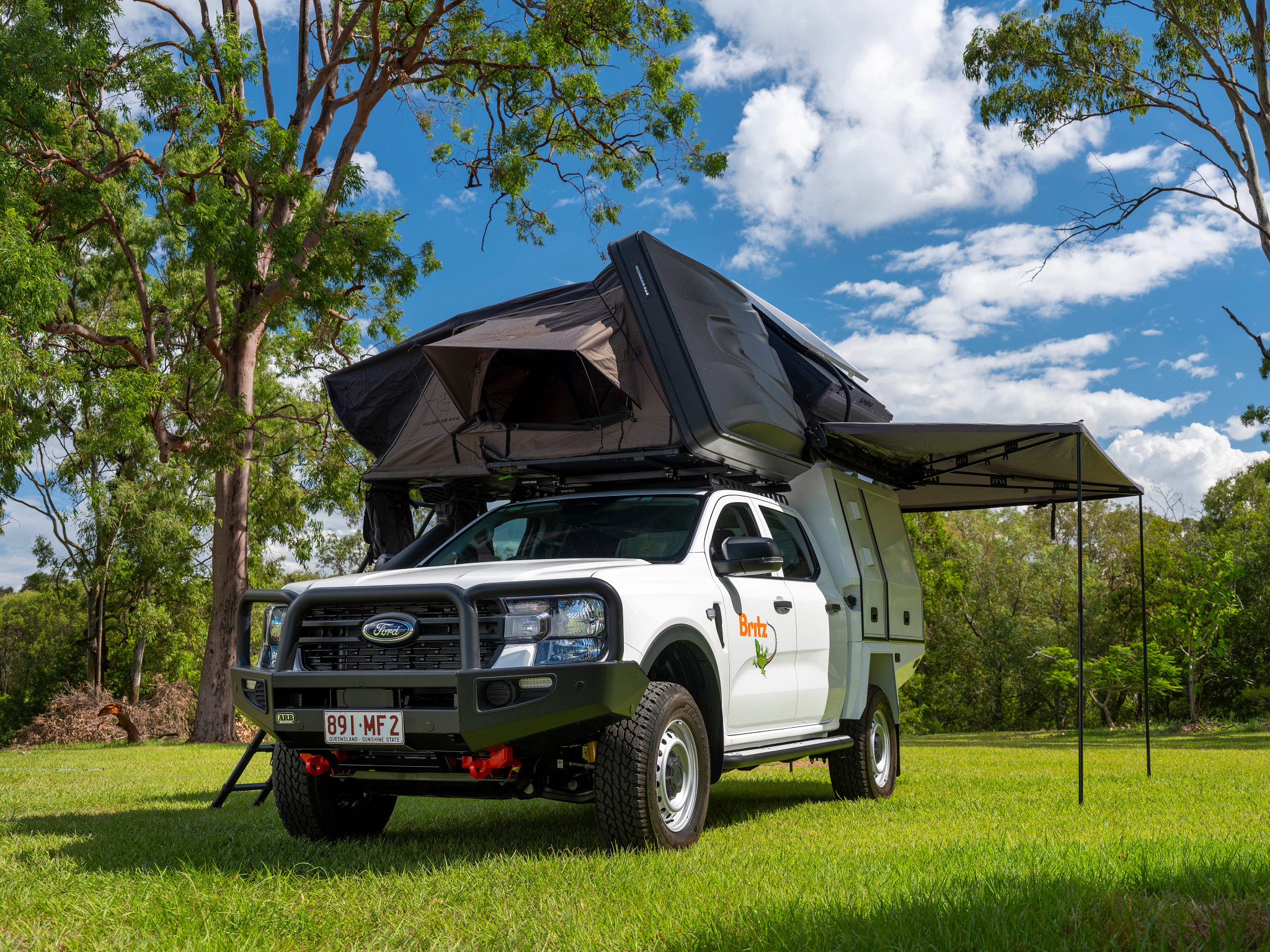 White Britz 4WD Ford campervan with an open rooftop tent and extended awning parked on a grassy field.