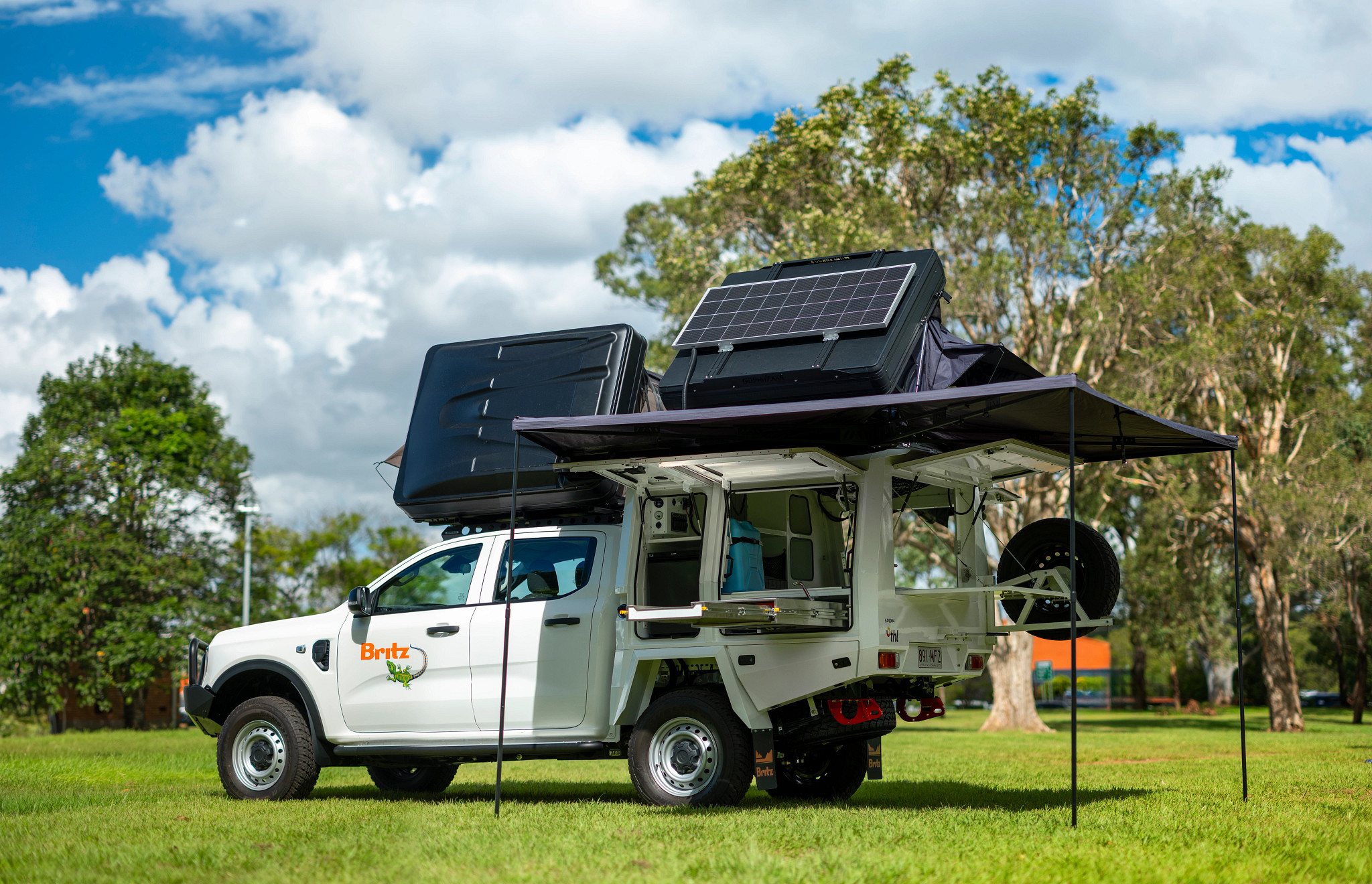 White Britz 4WD campervan with rooftop tents, solar panel, and side awning set up on a grassy field.