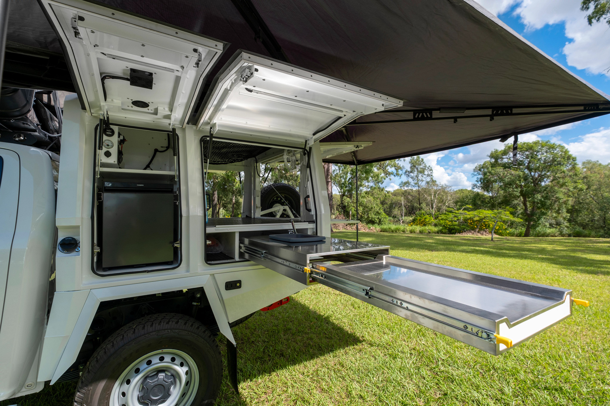 White campervan with open side canopy showing a built-in fridge and an extended stainless steel slide-out kitchen tray.