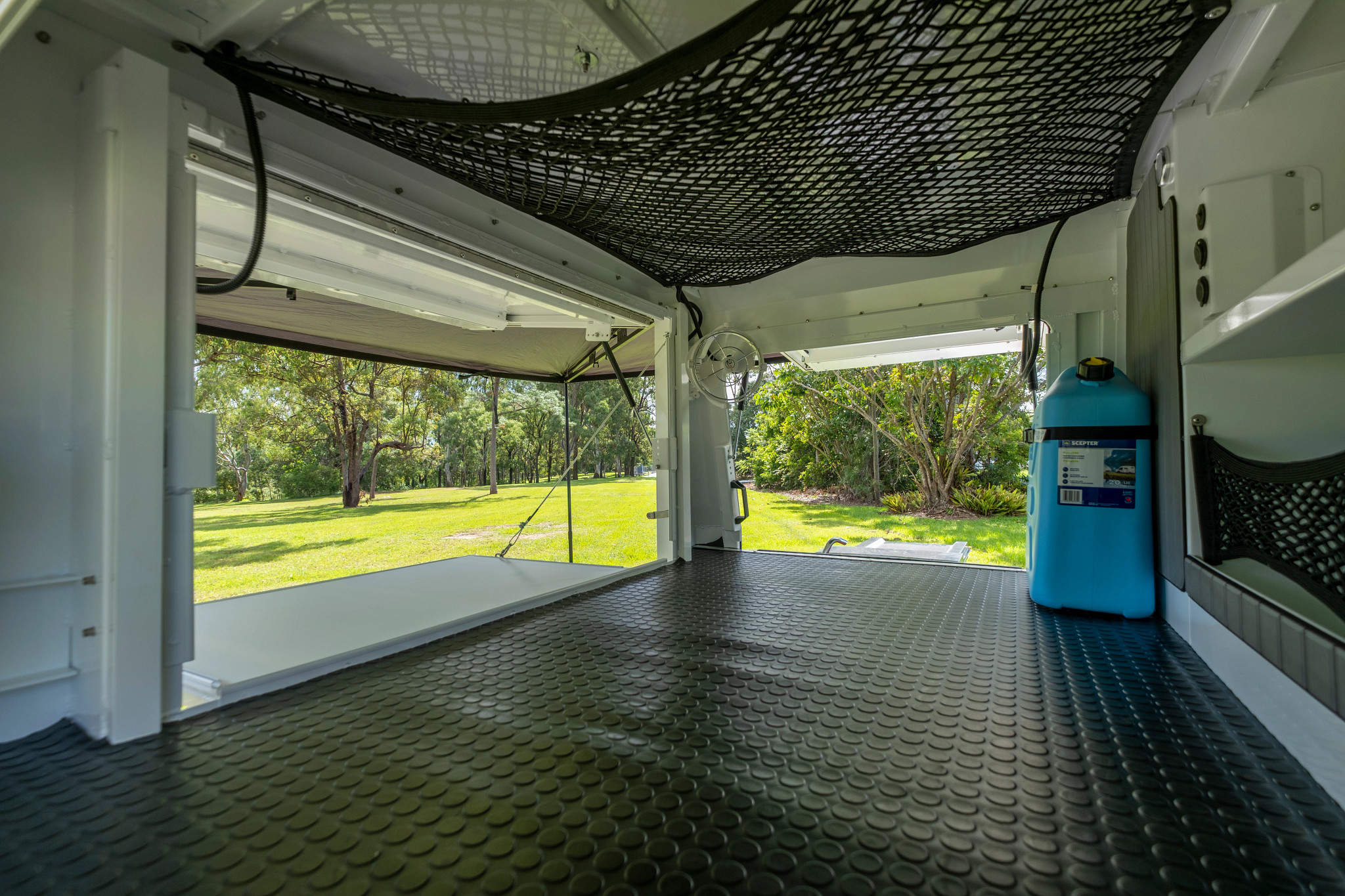 Inside a campervan with black coin flooring, a ceiling cargo net, and a blue jerry can, looking out at a green lawn.
