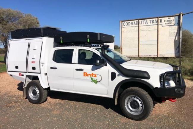 White Britz 4WD campervan with a rooftop tent parked next to a rustic Oodnadatta Track sign in the outback.