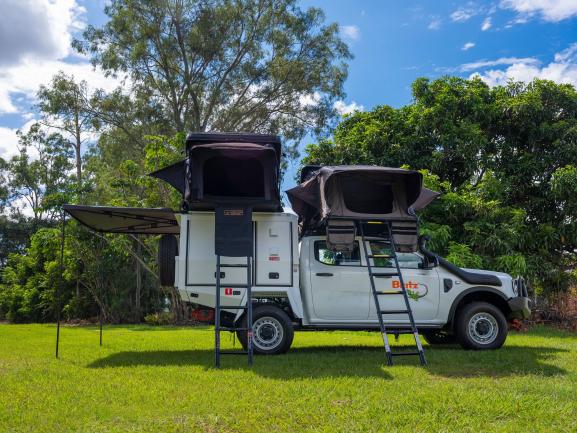 Britz 4WD campervan with two set-up rooftop tents, access ladders, and an extended side awning on a grassy field.