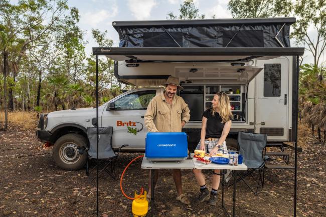 Man and woman prepare food on a portable table outside a white Britz 4WD campervan in the bush.