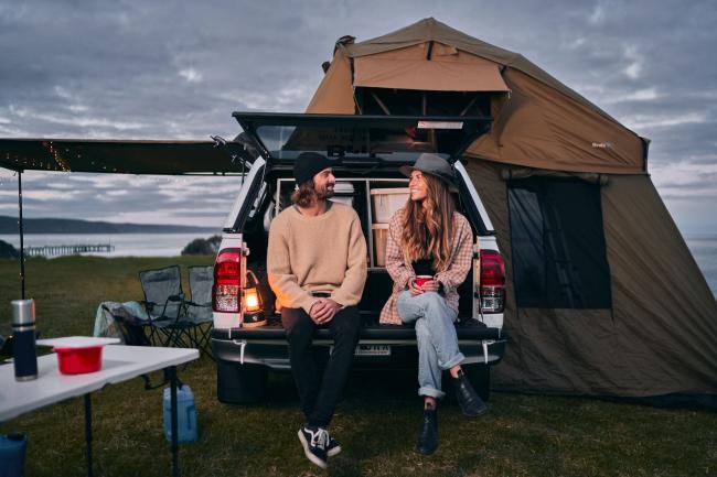 Couple sitting on the open tailgate of a white ute with a rooftop tent at a coastal campsite.