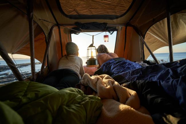Two people lying inside a campervan rooftop tent, looking out at the ocean at sunrise.