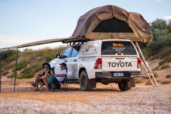 Couple sitting under the awning of a white Britz 4WD campervan with a pitched rooftop tent.