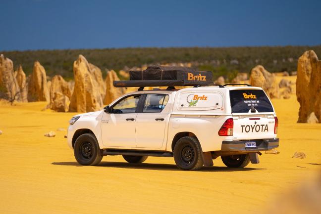 White Britz Toyota 4WD campervan with rooftop tent parked in a sandy desert with limestone pillars.