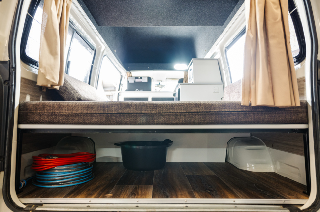 Campervan rear interior showing under-bed storage with a coiled hose and black tub on timber-look flooring.
