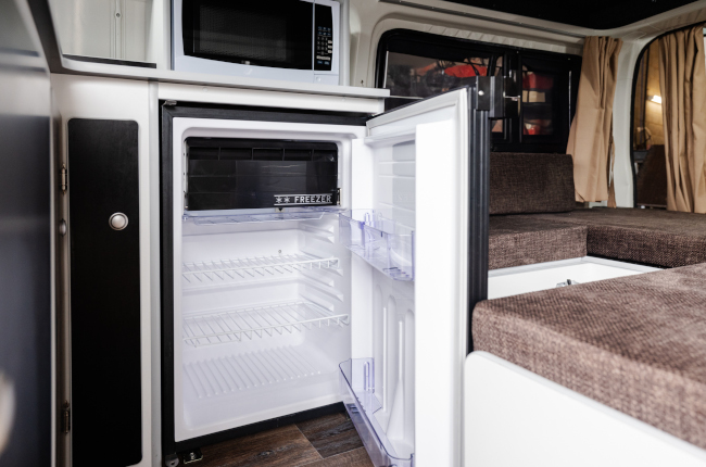Compact open fridge and microwave inside a campervan next to brown upholstered seating.