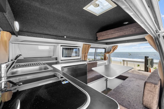 Campervan interior showing a kitchen sink and microwave beside a dining table and seating overlooking the ocean.