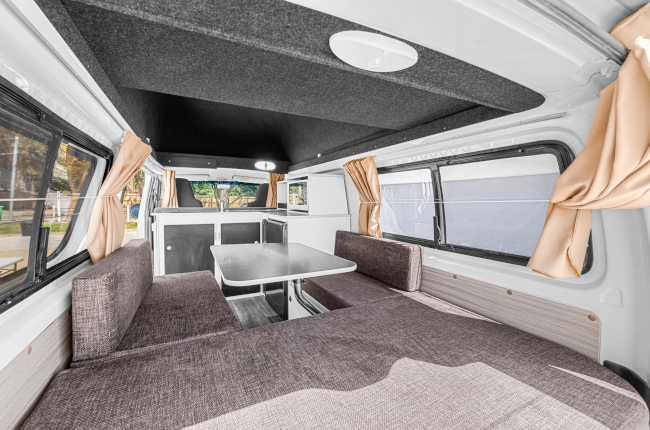 Campervan interior with grey fabric bench seats around a central table, white kitchen cabinets, and beige window curtains.