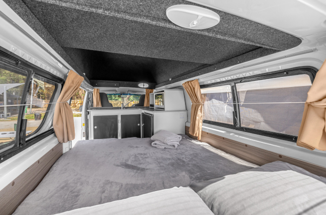 Campervan interior featuring a double bed with grey blankets, side windows with beige curtains, and a front kitchenette.