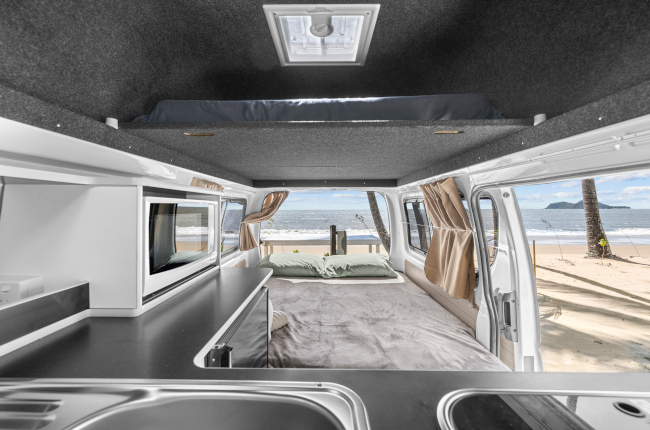 Campervan interior with a kitchenette, double bed, and loft bed looking out to a beach.