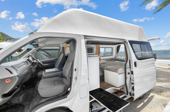 White high-top campervan with open doors showing grey front seats and a rear bed, parked near the ocean.