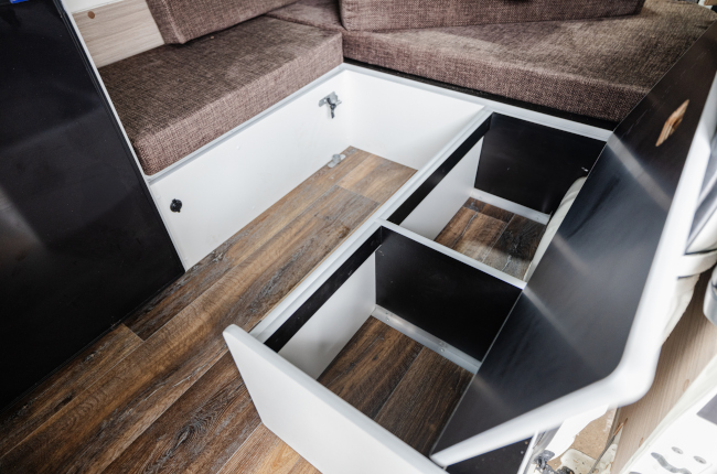Opened storage compartments beneath bench seats in a campervan with brown cushions and timber-look flooring.