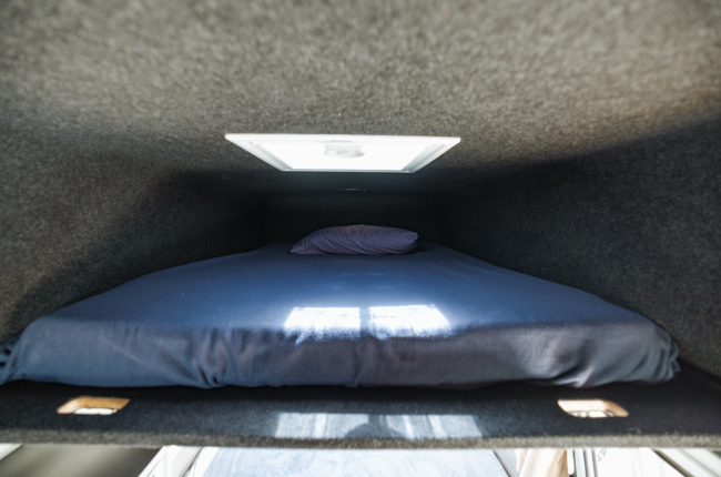 Grey carpeted campervan sleeping loft with a blue-sheeted mattress, a pillow, and a small overhead skylight.