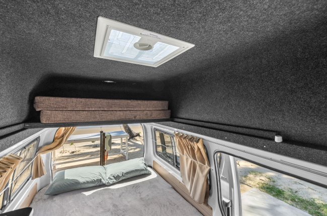 Campervan interior with a bed, green pillows, side windows, and a grey felt-lined high ceiling featuring a skylight.
