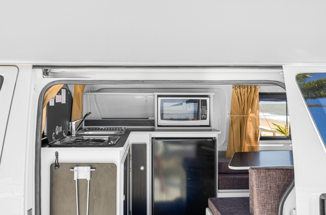 Compact campervan kitchen with a sink, microwave, mini fridge, dining table, and mustard-coloured curtains.