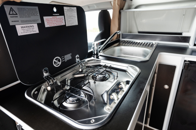 Stainless steel two-burner gas stove with an open lid next to a sink inside a campervan kitchen.