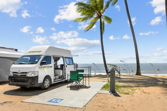 White Camperman campervan parked at a beachside campsite with folding chairs, palm trees, and ocean views.
