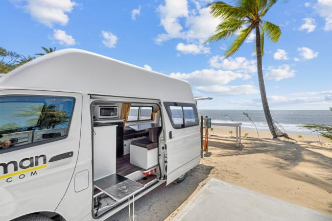 White campervan with an open side door showing the interior kitchen, parked on a sandy beach next to a palm tree.