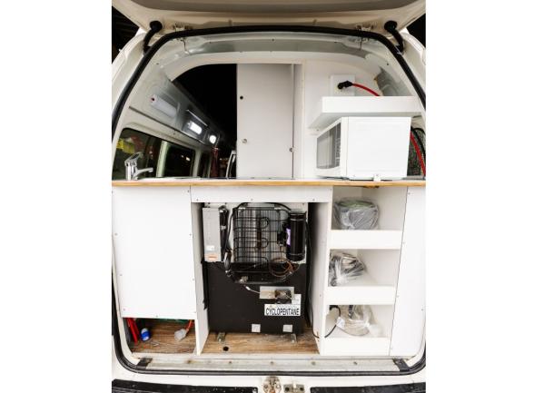 Converted campervan rear interior featuring a kitchen unit with a timber countertop, microwave, fridge, and shelves.