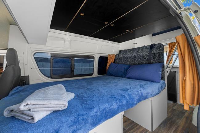 Campervan interior with a raised double bed made with a blue blanket, blue pillows, and folded grey towels.