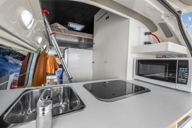 Compact campervan kitchen with a white benchtop, stainless steel sink, glass cooktop, and microwave.