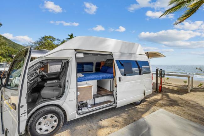 White campervan parked on a sandy beach with open doors showing a blue bed and interior.