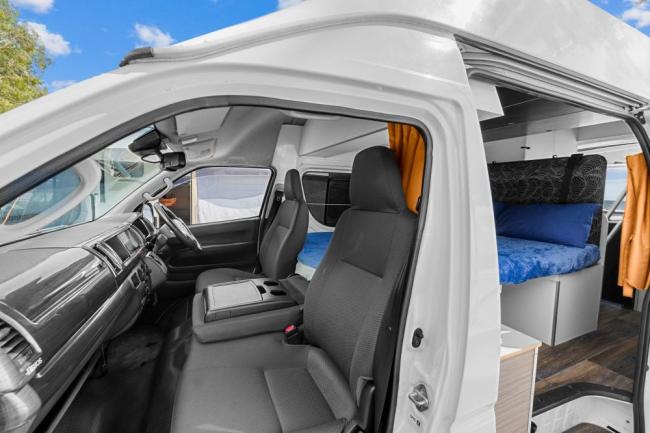 Campervan interior showing grey front seats and a rear cabin with a blue bed and orange curtains.