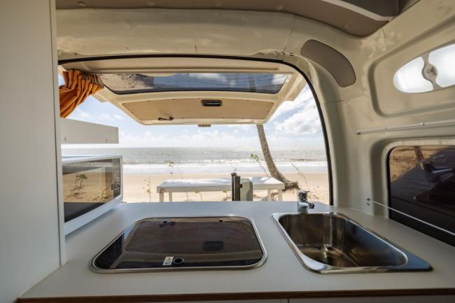 Campervan kitchen with sink and stove looking out the open rear door to a sandy beach and ocean.