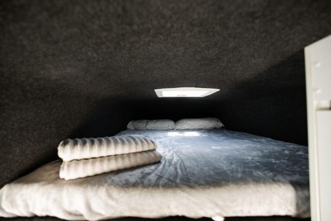 Campervan sleeping compartment with a low dark ceiling, a made bed, folded towels, and a small skylight.