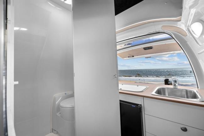 Campervan interior showing a toilet cubicle beside a kitchen counter with a sink overlooking an ocean view.