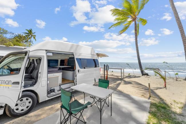 White campervan parked at a beach campsite with an outdoor table and green folding chairs by the ocean.