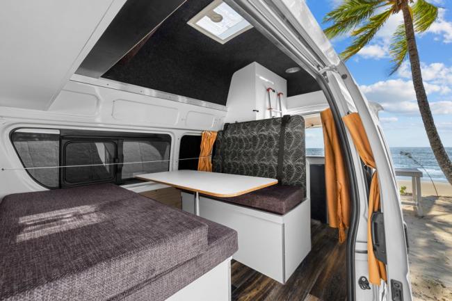 Campervan interior with a table and cushioned seats, looking out through the open door to a tropical beach.